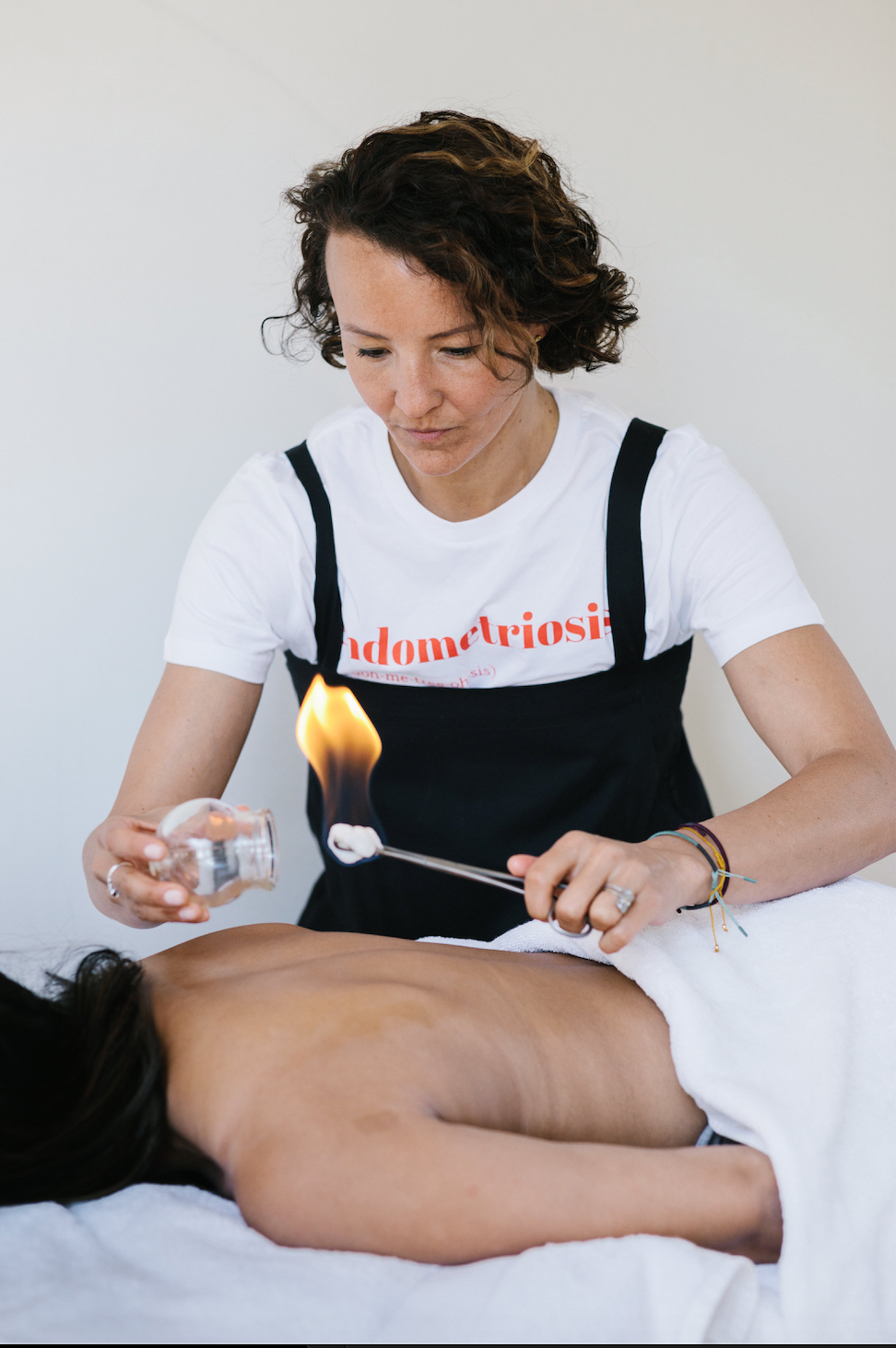 Person applying a product to another person's back with a lit candle, wearing a white t-shirt with red text.