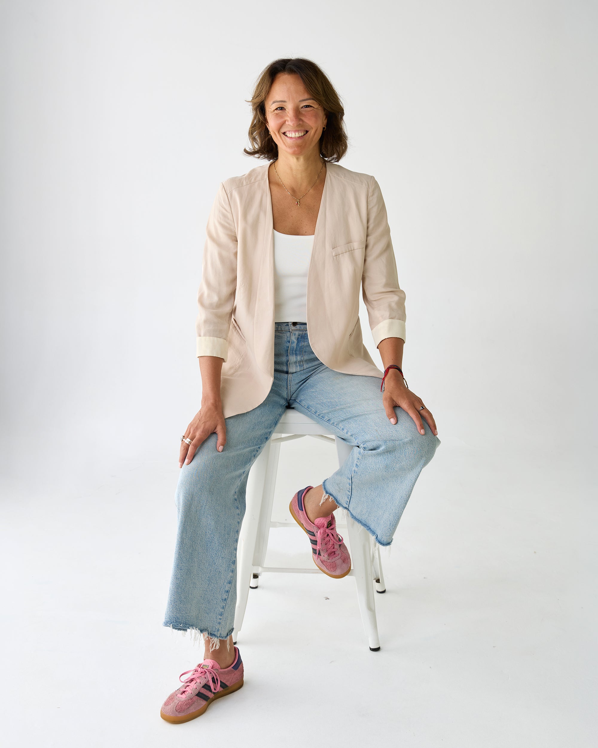 Woman sitting on a white stool wearing a beige cardigan, white top, blue jeans, and pink shoes 